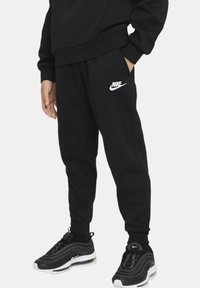 Pantalones de chándal negros con cintura elástica, bolsillos laterales y logotipo de Nike en blanco en la pierna izquierda, combinados con zapatillas negras.