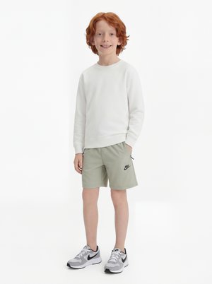 Enfant aux cheveux roux debout en souriant, portant un t-shirt blanc à manches longues, un short Nike gris clair et des baskets Nike grises sur fond blanc.