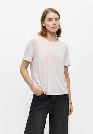 Kvinde med skulderlangt hår iført en lyserød kortærmet skjorte og mørke jeans, med den ene hånd i lommen, foran en ensfarvet hvid baggrund.