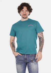 T-shirt verde acqua a maniche corte con la scritta 'PROPAGANDA' stampata in rosa. Texture morbida, scollatura rotonda, indossato con jeans in denim strappati e un accessorio in catena argentata.