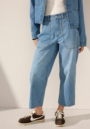 Personne portant un jean en denim bleu à fines rayures, une veste en denim assortie, un débardeur blanc et des baskets marron et blanches, debout sur un sol clair.