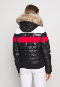 Vadderad svart jacka med vita och röda ränder, en huva fodrad med fuskpäls och ribbade mudd. Glansig yta med en kortad silhuett.