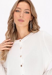 Bílý top s dlouhým rukávem, kulatým výstřihem, zlatými knoflíkovými akcenty a lehkou texturovanou látkou. Modelka má na sobě dlouhé visací náušnice.