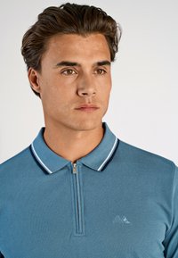 Męska koszulka polo w jasnoniebieskim kolorze, z zamkiem na szyi, granatowo-białym pasek przy kołnierzyku oraz delikatnym haftem logo na piersi.