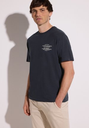 Homme portant un t-shirt bleu marine avec un texte blanc sur la poitrine, pantalon beige, debout contre un mur blanc avec une ombre projetée derrière.