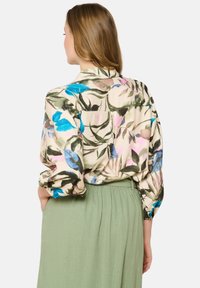 Blusa con estampado floral en tonos azul, rosa y verde, hecha de tela ligera; con mangas enrolladas y un bolsillo en la parte posterior.
