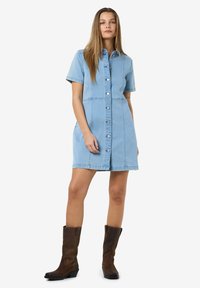 Vestido camiseiro de ganga azul claro com mangas curtas, fecho frontal com botões e costuras visíveis. Estilizado com botas castanhas até ao joelho.