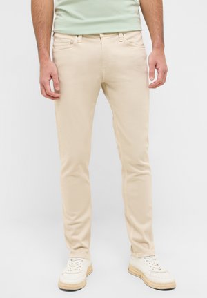 Beige denim jeans with a slim fit, five pockets, and minimal stitching, paired with white sneakers and a light green shirt.