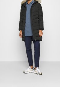 Zwarte gewatteerde jas met een met bont gevoerde capuchon, gecombineerd met een blauwe overhemd en marineblauwe broek, afgewerkt met witte en zilveren sneakers.