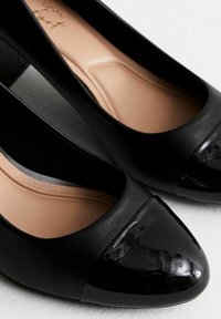 Zapatos de tacón de charol negro con cuerpo mate y puntera brillante. Cuenta con un interior liso y forma redondeada. Diseño elegante pero clásico.