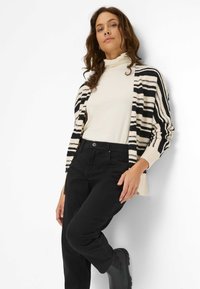 Stribet cardigan i sort og creme, tætsiddende silhuet, parret med en hvid turtleneck, sorte jeans og sorte ankelstøvler. Glat tekstur.