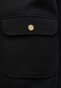 Μαύρο ύφασμα με τσέπη με καπάκι και κλείσιμο με χρυσό κουμπί. Το υλικό έχει υφή με ραβδώσεις, υπογραμμίζοντας την ανθεκτικότητα και το σχεδιασμό.