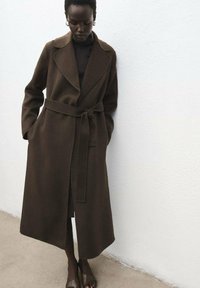 Abrigo de mezcla de lana marrón con cuello ancho, cinturón y bolsillos. El modelo está de pie frente a una pared blanca texturizada, llevando zapatos oscuros.