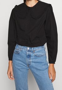 Blouse noire à manches longues avec un grand col en dentelle festonné, associée à un jean en denim bleu clair taille haute.