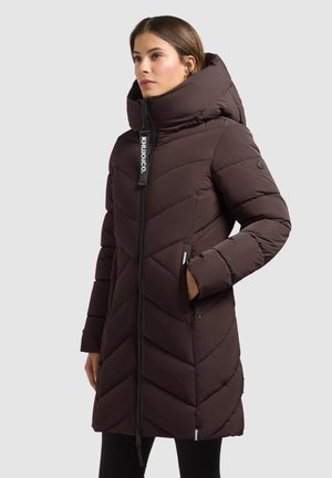 Mujer con abrigo de invierno acolchado de color marrón oscuro hasta la rodilla, con cuello alto, capucha y bolsillos laterales con cremallera, de pie con una mano en el bolsillo.