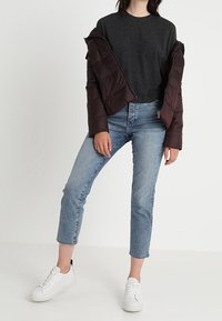 T-shirt gris foncé, veste matelassée marron, jean bleu clair et baskets blanches. La veste présente une texture matelassée et un design court.