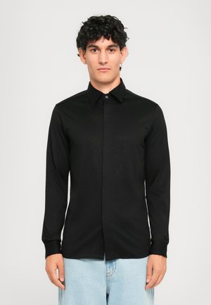 Joven con cabello oscuro y rizado que lleva una camisa negra de manga larga abotonada y jeans de color azul claro, de pie frente a un fondo blanco.