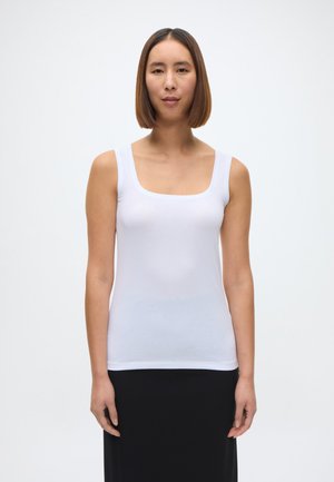 Fehér ujjatlan felső sima, könnyű anyagból készült. Szögletes dekoltázzsal és széles vállpántokkal rendelkezik, fekete szoknyával párosítva.