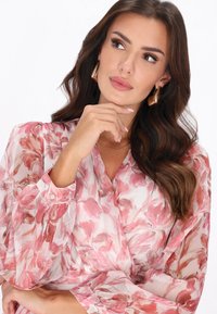 Blusa floral con tela suave y translúcida en tonos rosa y blanco. Presenta mangas largas y un diseño envuelto con detalles de botones sutiles.