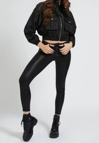 Mujer vistiendo una chaqueta corta negra, leggings negros de cintura alta y botas de combate negras con cordones, de pie contra un fondo claro y liso.