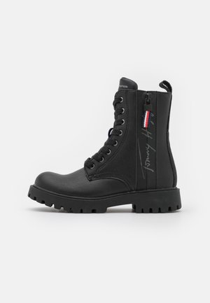 Sort snørestøvle i combat-stil med sidelås, tyk slidbane, trækklap og Tommy Hilfiger-branding på siden.
