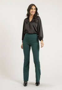 Blouse en satin noir avec un décolleté en V et des manches longues bouffantes, associée à un pantalon ajusté vert émeraude ; modèle portant des bottines noires.