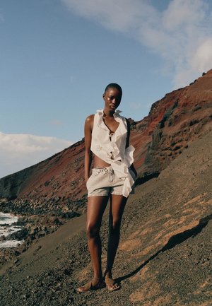 Žena u bijelom bez rukava, naboranom topu i traper kratkim hlačama stoji na kamenitom terenu s oceanom i liticama u pozadini pod plavim nebom.