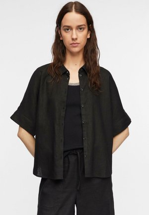 Młoda kobieta z długimi włosami, ubrana w luźną czarną koszulę na krótkim rękawie zapinaną na guziki, na czarnym topie i czarnych spodniach ze ściągaczem.