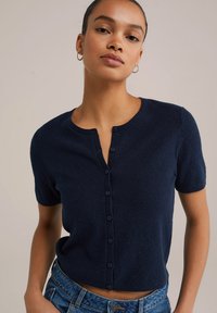 Tume sinine lühikeste varrukatega kudumiskardigan, millel on ümmargune kaelus ja nööpide sulgemine. Tekstureeritud kangas, kitsas siluett, sobib hästi denimpükstega.