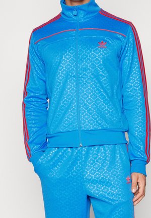 Mann trägt blauen Adidas-Trainingsanzug mit roten Streifen an Ärmeln und Hosen, Hände in den Taschen, mit kleinen Adidas-Logos auf Brust und Oberschenkel.