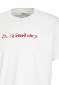 Witte t-shirt met gestileerde rode tekst die "Replay Speed Shop" leest, met een eenvoudige ronde halslijn.