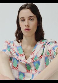 Młoda kobieta o długich brązowych włosach, ubrana w kolorową bluzkę z falbanami i abstrakcyjnymi zielonymi, czerwonymi i niebieskimi wzorami, o neutralnym wyrazie twarzy, na jednolitym tle.