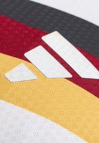 Gros plan de trois bandes blanches du logo Adidas sur un tissu avec des motifs de couleurs noir, rouge, jaune et blanc.