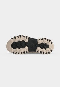 Semelle de chaussure noire et beige avec des rainures profondes conçues pour la traction et la durabilité, montrée sur un fond gris uni.