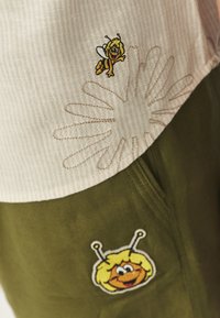 Katoenen outfit met een beige top met een bijborduring en een olijfgroene onderkant met een cartoon bijpatch en bloemenstiksel.