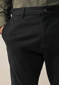 Schwarze Chinos aus Baumwollmischung, mit flachem Vorderteil, Seitentaschen und Knopfverschluss. Glatte Struktur mit minimalen Nähdetails.