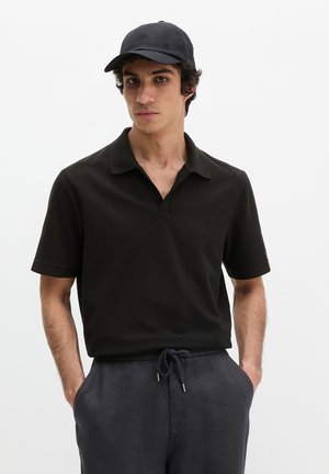 Jonge man met een zwart poloshirt, zwarte pet en donkere broek met trekkoord, staand met handen in de zakken tegen een effen achtergrond.