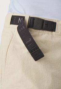 Närbild av beige byxor med ett svart flätat bälte som har en plastspänne med ett präglat varumärkeslogotyp.