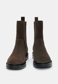 Botine Chelsea din suede maro, cu panouri elastice laterale, vârfuri rotunde și tălpi groase din cauciuc negru pentru aderență și durabilitate.
