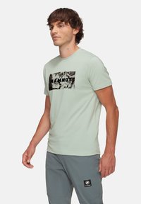 Ljusgrön t-shirt med kort ärm som har en svart rektangulär grafik med texten "MAMMUT." Tyget är mjukt och lätt.