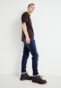 Dark brown T-shirt paired with blue jeans. Brown lace-up boots feature yellow stitching and a textured finish. Simple, casual outfit.