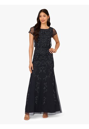 Vrouw die een zwarte avondjurk draagt die tot op de grond reikt, met doorschijnende korte mouwen met borduursels en decoratieve bloemmotieven, staand met één hand op haar heup.