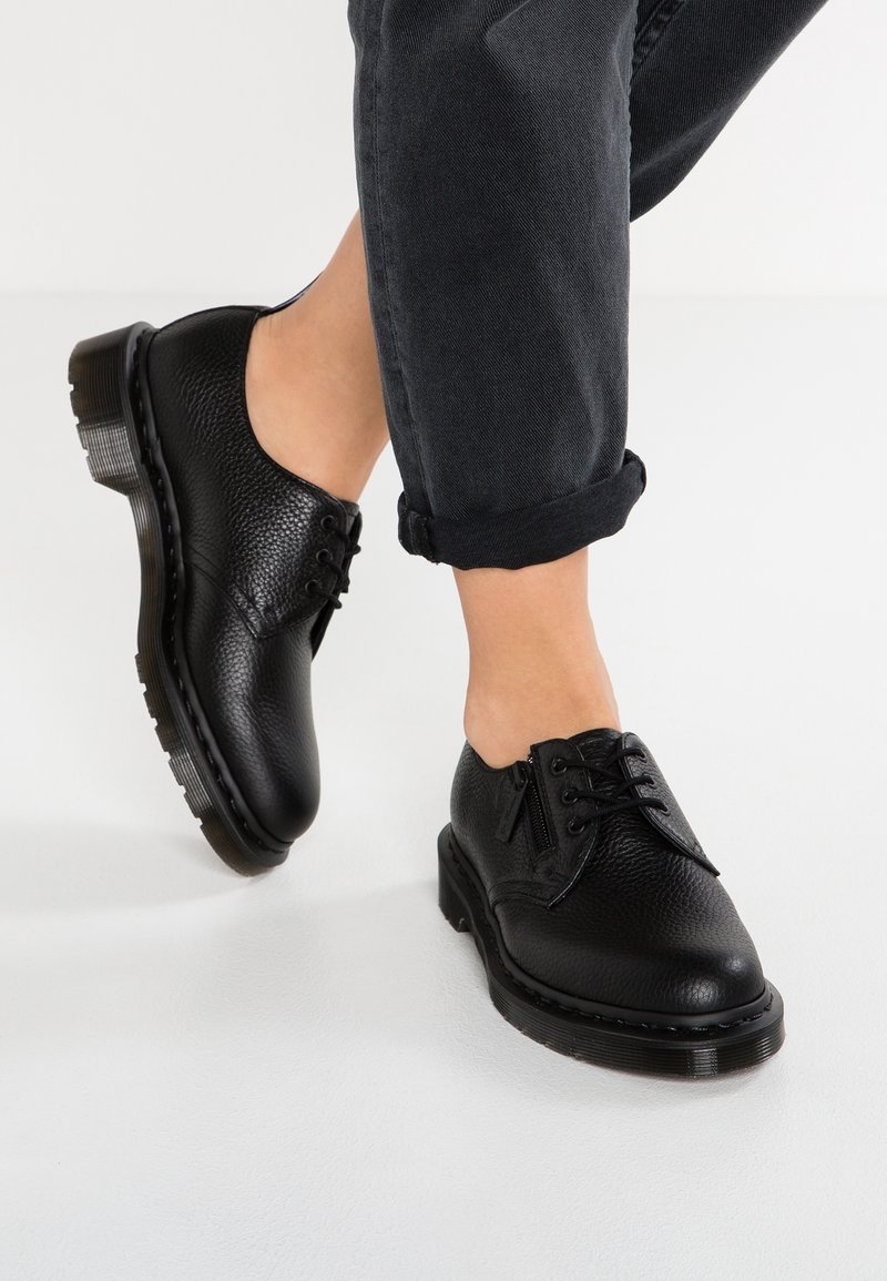 Chaussures à lacets en cuir noir avec finition texturée, bout rond et semelle épaisse en caoutchouc. Portées avec un pantalon foncé retroussé, mettant en valeur le design de la chaussure.