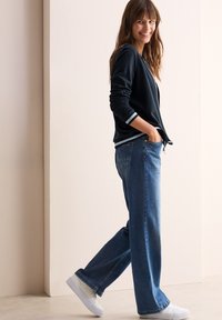Mörkblå cardigan med vita ränder vid ärmarna, tillsammans med löst sittande blå denimjeans och vita sneakers, mot en neutral bakgrund.