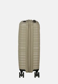Valigia rigida beige, con superficie testurizzata e scanalata, chiusura a doppio zip e quattro ruote nere per la mobilità.