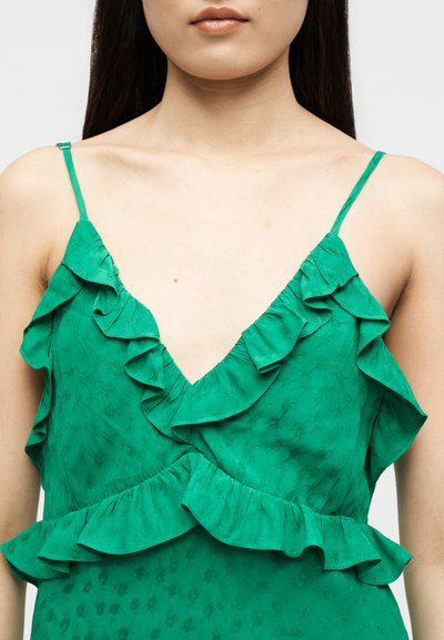 Νέος/Νέα που φοράει ένα πράσινο, αμάνικο φόρεμα με βολάν στις άκρες και λεπτές τιράντες, με εμφανή το λαιμό και το πάνω μέρος του στήθους.