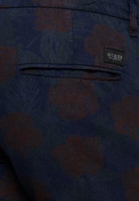 Tessuto blu navy con un motivo floreale marrone scuro. Presenta una tasca posteriore con etichetta GUESS. Superficie testurizzata.