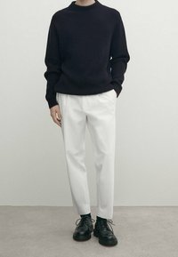 Personne portant un pull en tricot noir, un pantalon blanc et des chaussures à lacets noires, debout devant un fond clair uni.