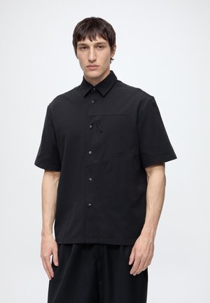 Un homme se tient face à nous, portant une chemise noire à manches courtes avec des boutons et une poche poitrine zippée sur un fond clair uni.