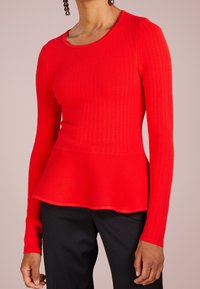 Rotes geripptes Langarmoberteil mit einem Peplumsaum, Rundhals und texturiertem Stoff. Kombiniert mit schwarzen Hosen.